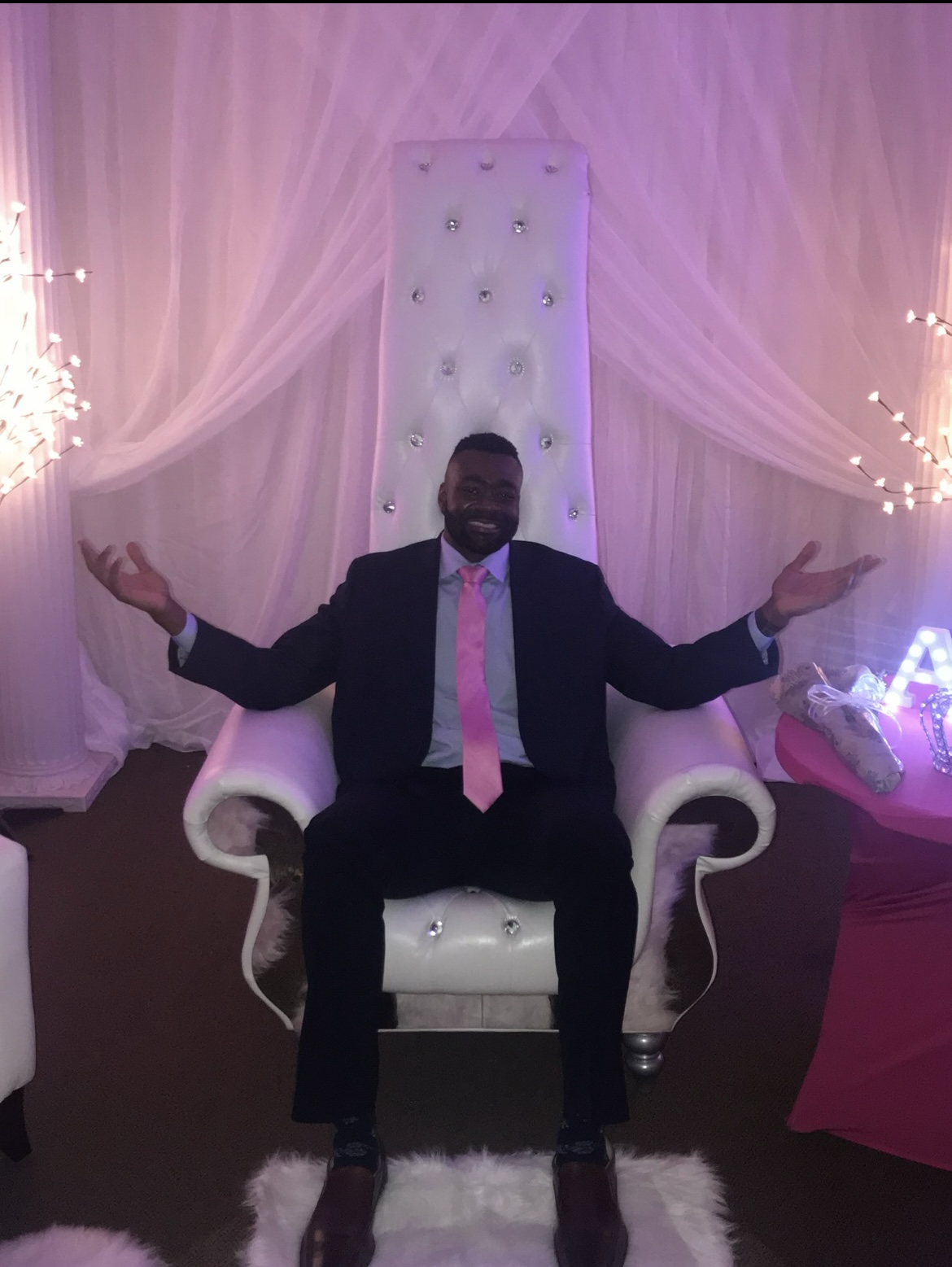 Guest seated on white tufted throne chair with pink uplighting and draped backdrop